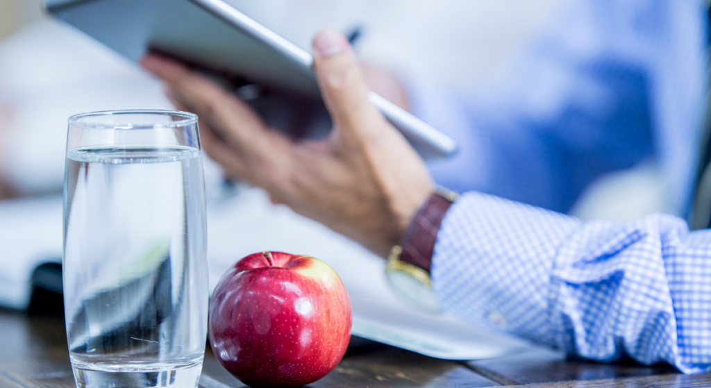 Ein Apfel liegt neben einem Glass Wasser. Im Hintergrund ist eine Person mit Tablet zu erkennen.